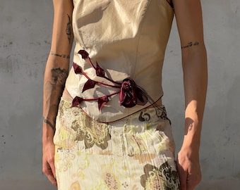 May include: A strapless beige corset top with a sweetheart neckline and dark red piping, featuring a dark red floral appliqué on the side. Paired with a cream-colored midi skirt decorated with a brown, yellow, and pink paisley-inspired pattern.