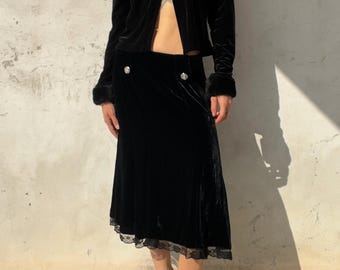 Könnte beinhalten: Eine langärmelige Jacke aus schwarzem Samt mit Kragen und Manschetten aus Kunstfell, kombiniert mit einem passenden Midirock aus schwarzem Samt mit zwei silberfarbenen Knöpfen an der Taille und schwarzer Spitzenborte am Saum.