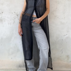 May include: A sleeveless, sheer black button-down duster vest worn over a gray tank top and light gray denim jeans, paired with black strappy sandals. The long, translucent garment features a classic collar and a full-length front button closure.