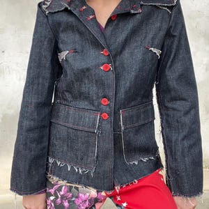 Puede incluir: Una chaqueta de mezclilla oscura con cuello de muesca, mangas largas y cierre frontal de cinco botones de color rojo brillante. La chaqueta tiene dos bolsillos delanteros con solapa, bordes deshilachados en costuras y dobladillo, y costuras rojas.