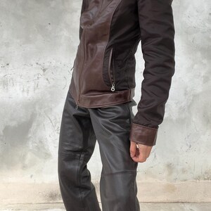 May include: A brown leather jacket with a collared neckline and a zipper closure. The jacket has brown leather panels and dark brown fabric sleeves. The outfit includes dark brown leather pants.