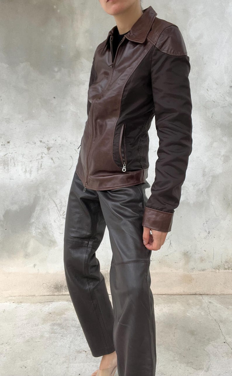 May include: A brown leather jacket with a collared neckline and a zipper closure. The jacket has brown leather panels and dark brown fabric sleeves. The outfit includes dark brown leather pants.