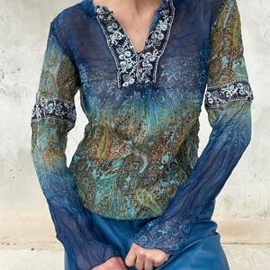 Può includere: Una camicetta a maniche lunghe con motivo paisley blu e verde su tessuto trasparente e stropicciato. Il capo presenta uno scollo a V con un bordo ricamato floreale bianco e nero che corre lungo il centro e sulle maniche superiori.