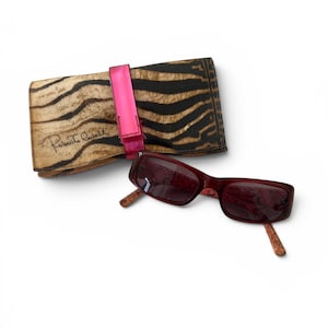 May include: A pair of dark red rectangular sunglasses with patterned temples and a matching rectangular folding case featuring a black and tan zebra print, a bright pink strap, and the Roberto Cavalli brand name printed on the front.