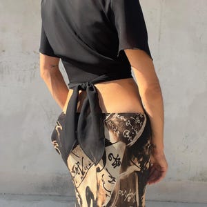 May include: A black short-sleeved wrap top with a tie closure at the lower back, paired with a long skirt featuring a black and tan abstract print with handwritten-style script text throughout the fabric, creating a layered and patterned appearance.