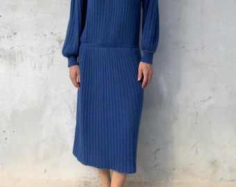 Könnte beinhalten: Ein blaues, geripptes Strickkleid in Midilänge mit hohem Ausschnitt und langen Ärmeln. Der obere Brustbereich hat eine kontrastierende dunkelblaue Passe, die mit kleinen silbernen Nieten und einer goldenen Perlenborte verziert ist.