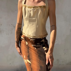 May include: A cream-colored corset top featuring structured vertical seams, thick shoulder straps, and gold beaded trim along the neckline and hem. Paired with a brown and black midi skirt decorated with a light-colored leaf pattern throughout.