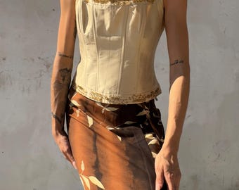 May include: A cream-colored corset top featuring structured vertical seams, thick shoulder straps, and gold beaded trim along the neckline and hem. Paired with a brown and black midi skirt decorated with a light-colored leaf pattern throughout.