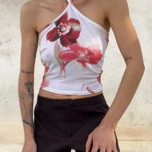 May include: A white halter top featuring a large red and pink floral print. The garment has a high neckline with thin straps that wrap around the neck, a sleeveless design, and a cropped length that sits above the waistline of the dark trousers.
