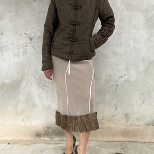 May include: A dark olive green quilted jacket with a high collar and three decorative toggle closures, paired with a beige midi skirt featuring vertical white piping and a pleated dark olive green hem, worn with dark olive green pointed-toe heels.