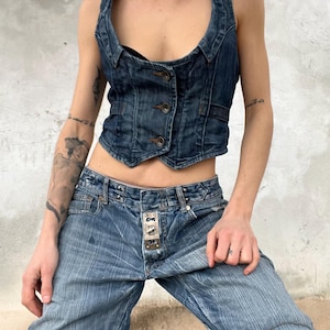May include: A dark blue denim vest featuring a scoop neckline, three front buttons, and a cropped hemline, paired with light blue denim jeans that have a distinct vertical striped texture and a prominent metal button fly closure detail.