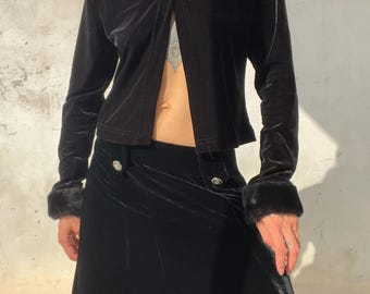 Könnte beinhalten: Eine langärmelige Jacke aus schwarzem Samt mit einem Kragen und Ärmelbündchen aus Kunstfell, kombiniert mit einem passenden Rock aus schwarzem Samt, der zwei silberfarbene Zierknöpfe an der Taille aufweist. Die Jacke ist vorne offen.