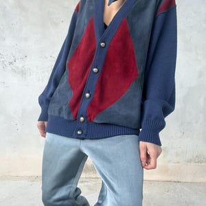 Op de afbeelding: Een vintage vest met een colorblock-ontwerp. De trui heeft een marineblauwe basis met rode en grijze suède ruitvormen op de voorkant. De mouwen en de boorden zijn marineblauw. Het vest heeft zilveren knopen.