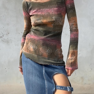 May include: A long-sleeve top featuring a dark olive, orange, and pink paisley-inspired pattern with a wide neckline and lettuce-edge hem, paired with a blue denim midi skirt that has a high side slit secured by a metal buckle strap.