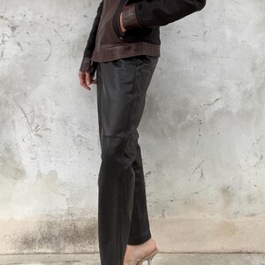 May include: A brown and black leather jacket with a collared neckline and a zipper closure. The jacket is paired with black leather pants and clear heeled sandals. The outfit is a modern take on a classic style.