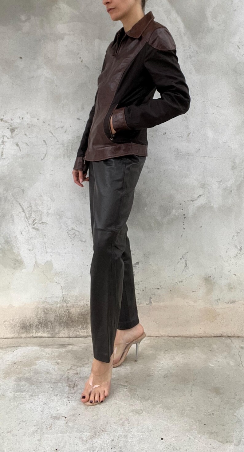 May include: A brown and black leather jacket with a collared neckline and a zipper closure. The jacket is paired with black leather pants and clear heeled sandals. The outfit is a modern take on a classic style.