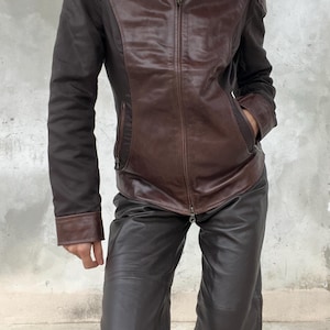 May include: A brown leather jacket with a zip-up front and a collared neckline. The jacket has contrasting dark brown sleeves and is paired with matching brown leather pants. The outfit is a vintage style.