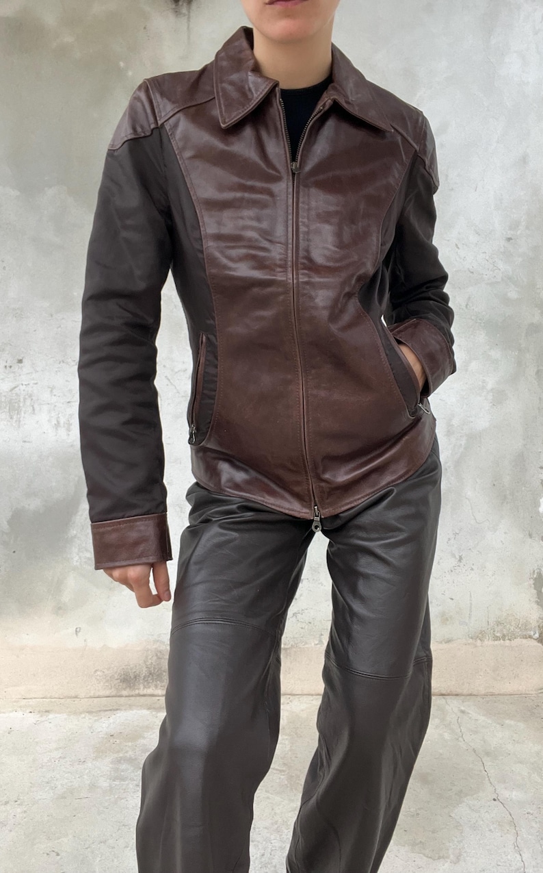 May include: A brown leather jacket with a zip-up front and a collared neckline. The jacket has contrasting dark brown sleeves and is paired with matching brown leather pants. The outfit is a vintage style.