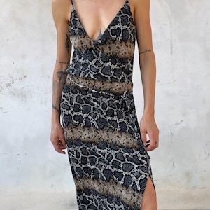 May include: A sleeveless midi dress featuring a snakeskin pattern in shades of black, gray, and tan. The dress has a V-neckline, thin spaghetti straps, a fitted bodice, and a wrap-style skirt with a side slit and a tie detail at the waist.