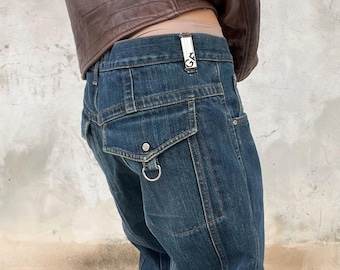 Könnte beinhalten: Dunkelblaue Jeans aus Denim mit einem silbernen Metallclip am Bund mit den Buchstaben GA, einer Pattentasche an der Hüfte und einem silbernen D-Ring unter der Tasche. Die Jeans wird mit einer braunen Lederjacke kombiniert.