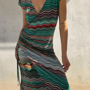 Può includere: Un abito a portafoglio di lunghezza midi caratterizzato da un vivace motivo a chevron ondulato multicolore nei toni del verde ottanio, nero, rosso e arancione. L'abito presenta maniche corte, un profondo scollo a V e un laccio laterale.