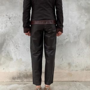 May include: A dark brown jacket with brown leather accents on the shoulders, collar, and cuffs. The jacket is paired with matching dark brown leather pants. The outfit is completed with clear heeled sandals.