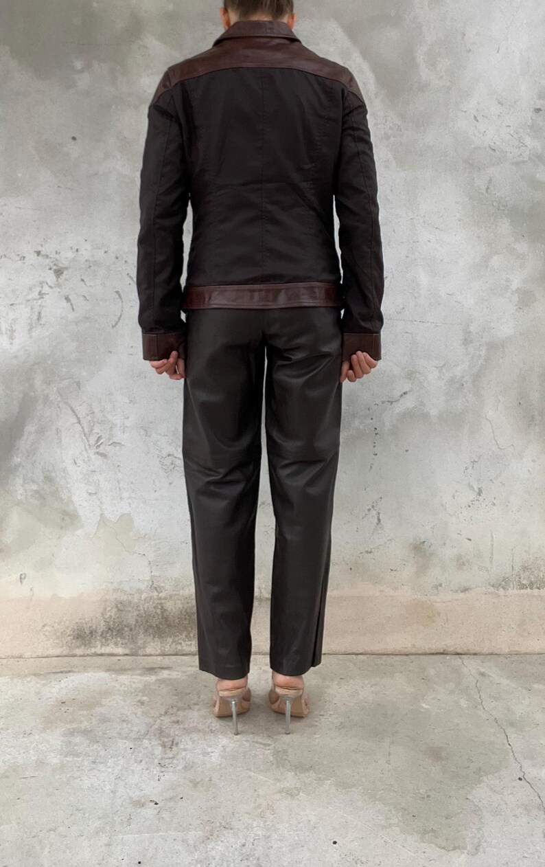 May include: A dark brown jacket with brown leather accents on the shoulders, collar, and cuffs. The jacket is paired with matching dark brown leather pants. The outfit is completed with clear heeled sandals.