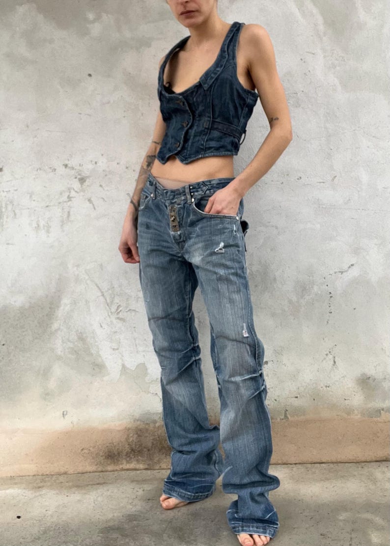 May include: A dark blue denim vest with a button-up front and a cropped hem paired with medium-wash blue denim jeans featuring a button-fly closure, distressed detailing, and a straight-leg fit that pools slightly at the bottom hem.