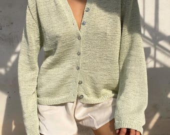 Könnte beinhalten: Eine hell salbeigrüne Strickjacke mit V-Ausschnitt, langen Ärmeln und einer Knopfleiste auf der Vorderseite mit kleinen runden Knöpfen. Das Kleidungsstück hat ein strukturiertes Strickmuster und wird mit schlichten, glatten Shorts kombiniert.