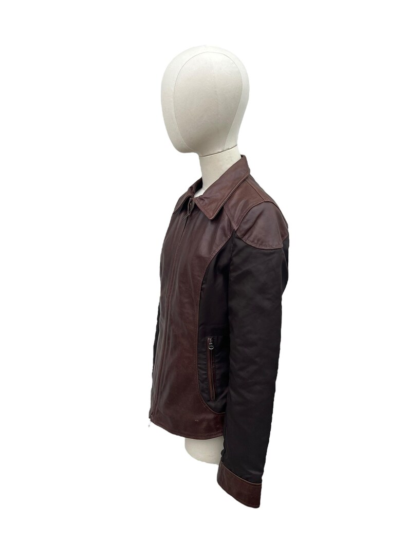 May include: A brown and black leather jacket with a collared neckline and a zipper closure. The jacket has a contrasting black panel on the sleeves and a small pocket on the chest. The jacket is displayed on a mannequin.