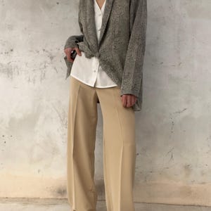 Peut inclure: Une femme porte un blazer gris à motif peau de serpent sur une chemise blanche boutonnée et un pantalon large beige. La tenue est dans une palette de couleurs neutres, le blazer étant le point central. Le pantalon est long.