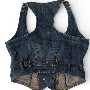 May include: A dark blue denim vest featuring a racerback design, a waist belt with a brass-toned metal buckle, and a pointed hemline. The interior lining and edges are finished with a colorful floral pattern featuring small pink and green flowers.