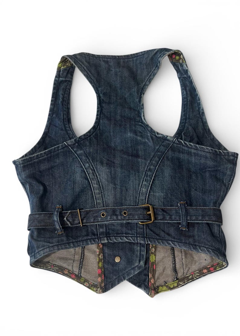 May include: A dark blue denim vest featuring a racerback design, a waist belt with a brass-toned metal buckle, and a pointed hemline. The interior lining and edges are finished with a colorful floral pattern featuring small pink and green flowers.