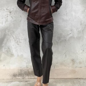 May include: A brown leather jacket with black fabric panels and a zipper closure. The jacket is paired with black leather pants. The person is wearing clear sandals. The outfit is set against a gray concrete wall.