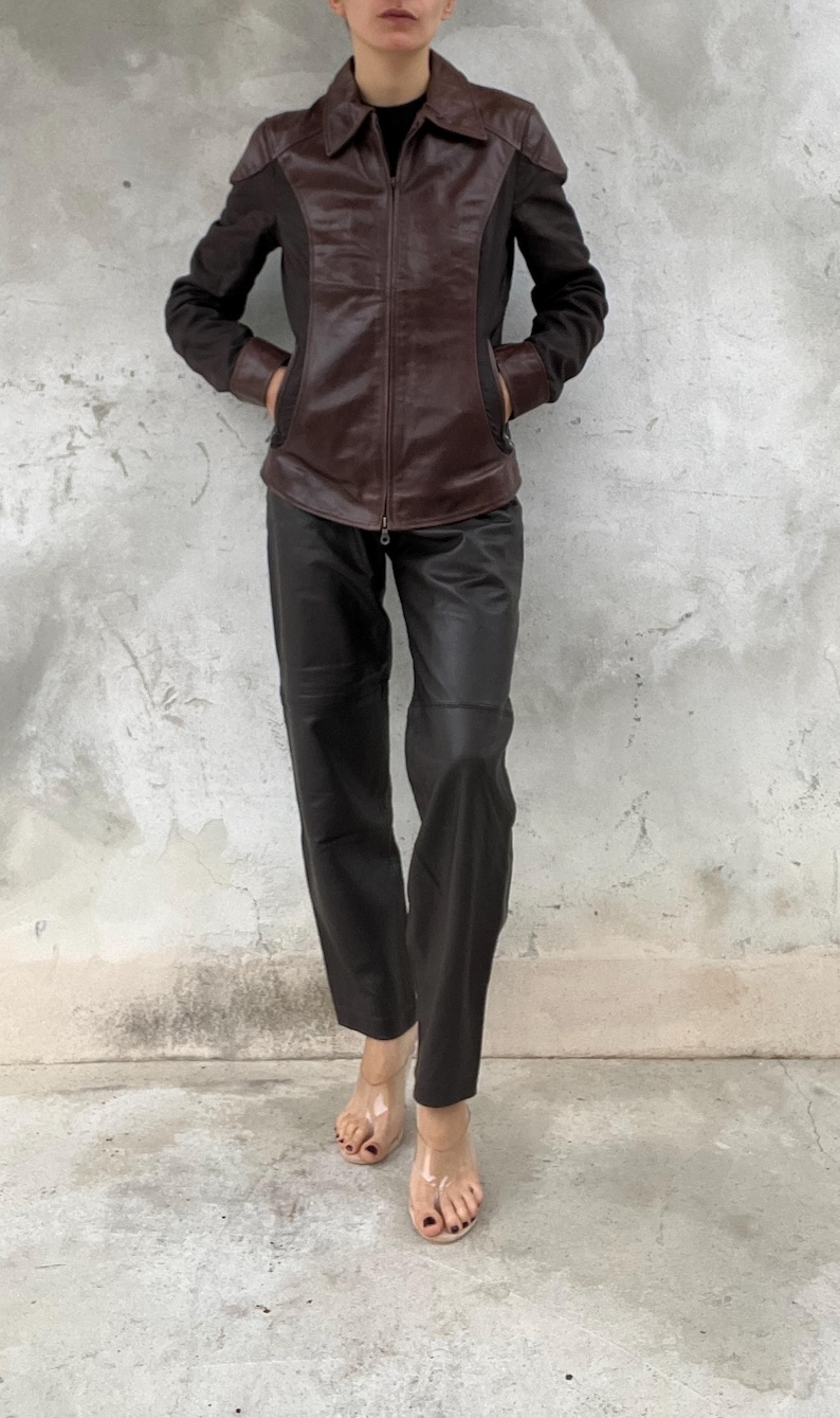 May include: A brown leather jacket with black fabric panels and a zipper closure. The jacket is paired with black leather pants. The person is wearing clear sandals. The outfit is set against a gray concrete wall.
