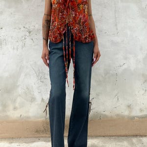Op de afbeelding: Een mouwloze top met V-hals en een paisley patroon in rood-, oranje- en geeltinten, gecombineerd met een donkerblauwe flared jeans. De jeans heeft decoratieve bandjes aan de zijkanten, wat een vintage-geïnspireerde outfit creëert.
