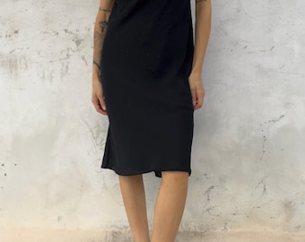 Könnte beinhalten: Ein schwarzes, knielanges Kleid mit Wasserfallausschnitt und dünnen Spaghettiträgern. Der Stoff weist durchgehend eine subtil texturierte Optik auf, die vom Ausschnitt bis zum Saum einen einheitlich dunklen Farbton erzeugt.