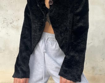 May include: A black faux fur jacket with a collared neckline and button closure. The jacket has long sleeves and a textured appearance. The model is wearing gray sweatpants. The jacket is a vintage style.