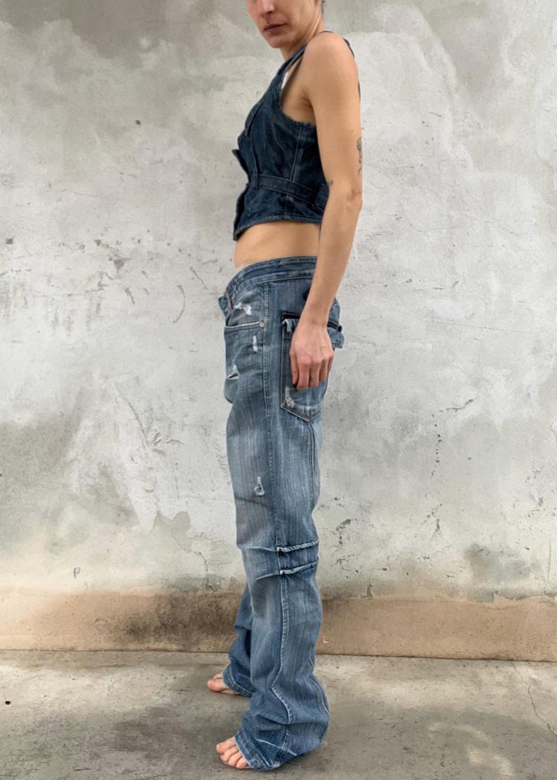 May include: A dark blue denim vest with a button-up front and a cropped hem paired with light blue distressed denim jeans featuring a low-rise waist, multiple pockets, and horizontal seam detailing along the legs in a relaxed, straight-leg fit.