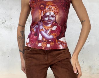 Könnte beinhalten: Ein ärmelloses Tanktop mit einem lebendigen, detaillierten Grafikdruck der hinduistischen Gottheit Krishna, die Flöte spielt, in den Farben Dunkelrot, Orange und Gold. Das Oberteil hat dünne Träger und einen Rundhalsausschnitt, kombiniert mit einer braunen Hose.