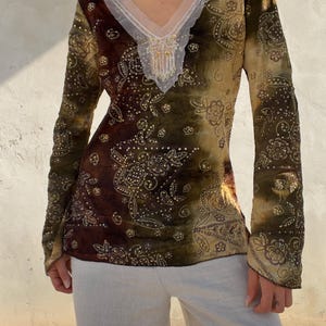 May include: A long-sleeved top featuring a deep V-neckline with white sheer trim and intricate beadwork. The fabric displays a tie-dye effect in shades of brown, olive green, and tan, adorned with a gold floral pattern and scattered sequins.