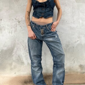 May include: A dark blue denim vest with a scoop neckline and three front buttons, paired with matching dark blue denim jeans featuring a distressed finish, a straight-leg cut, and a prominent metal button closure at the waistline.