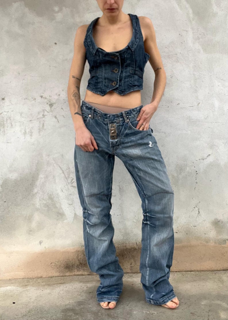 May include: A dark blue denim vest with a scoop neckline and three front buttons, paired with matching dark blue denim jeans featuring a distressed finish, a straight-leg cut, and a prominent metal button closure at the waistline.