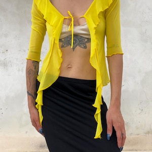 May include: A bright yellow sheer mesh cardigan featuring three-quarter length sleeves, a ruffled trim along the front opening, and a thin tie closure at the neckline. The garment is worn over a beige top and paired with a black skirt.