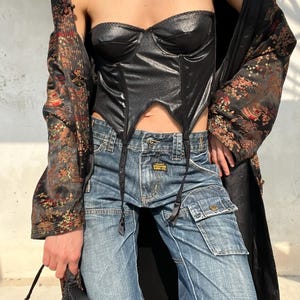 Peut inclure: Un haut corset noir sans bretelles à la finition texturée et à l'ourlet pointu, porté avec un jean cargo bleu en denim doté de poches latérales et une veste noire à manches longues à motifs, accompagné d'un petit sac à main noir perlé.