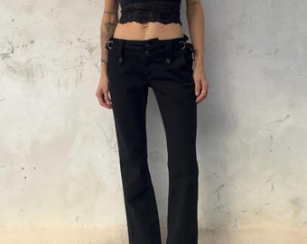 Könnte beinhalten: Ein schwarzes ärmelloses bauchfreies Oberteil mit Spitzenbesatz und Spaghettiträgern, kombiniert mit einer schwarzen tief sitzenden Hose mit Knopfverschluss, Gürtelschlaufen und D-Ring-Metallbeschlägen.
