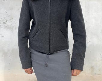 Könnte beinhalten: Eine anthrazitfarbene Reißverschlussjacke mit dunkelbraunem Kunstfellkragen und zwei Vordertaschen, kombiniert mit einem langen, eng anliegenden grauen Rock, der eine Kordelzugtaille mit kleinen schwarzen Stoppern in der Mitte aufweist.