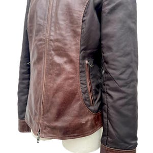May include: A brown leather jacket with a zip-up front and a collared neckline. The jacket features dark brown sleeves and a pocket on the front. The leather has a distressed appearance. The jacket is displayed on a white mannequin.