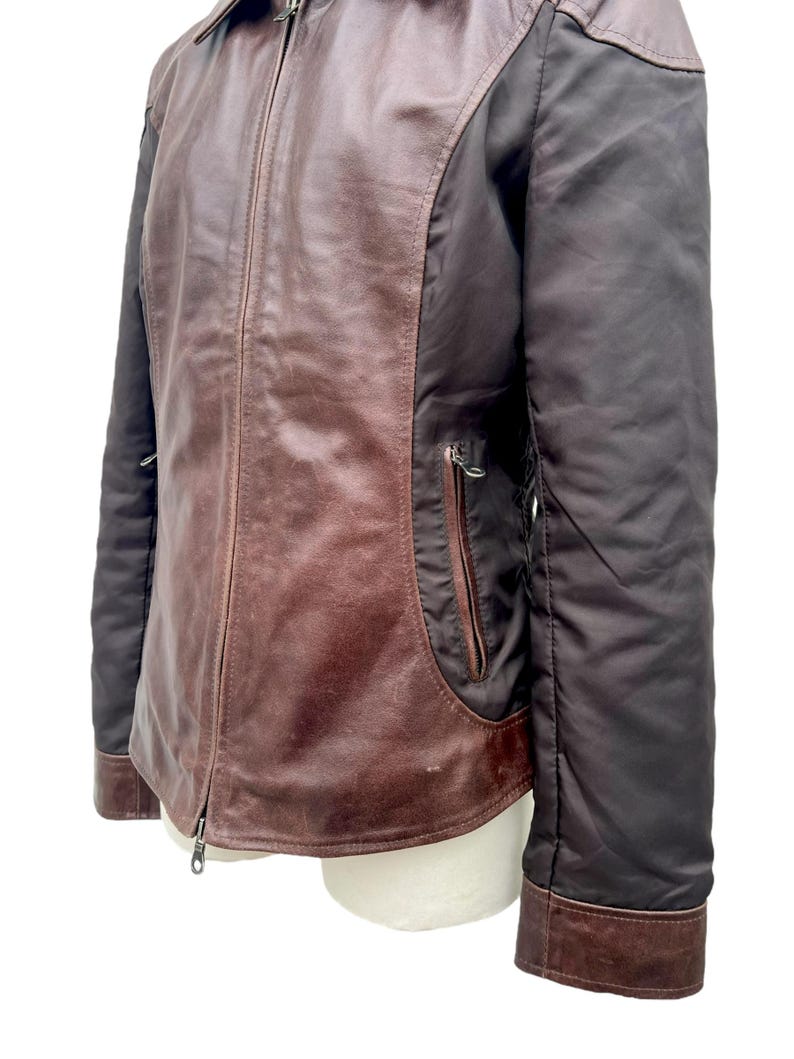 May include: A brown leather jacket with a zip-up front and a collared neckline. The jacket features dark brown sleeves and a pocket on the front. The leather has a distressed appearance. The jacket is displayed on a white mannequin.