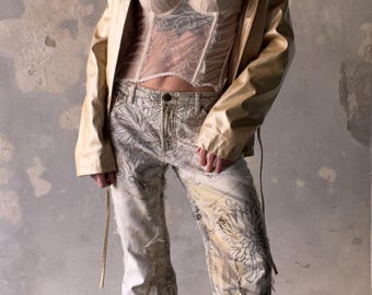 May include: A cream-colored corset with lace detailing is paired with a gold jacket and off-white jeans featuring a black floral design. The outfit is a blend of textures and styles, creating a unique fashion statement.
